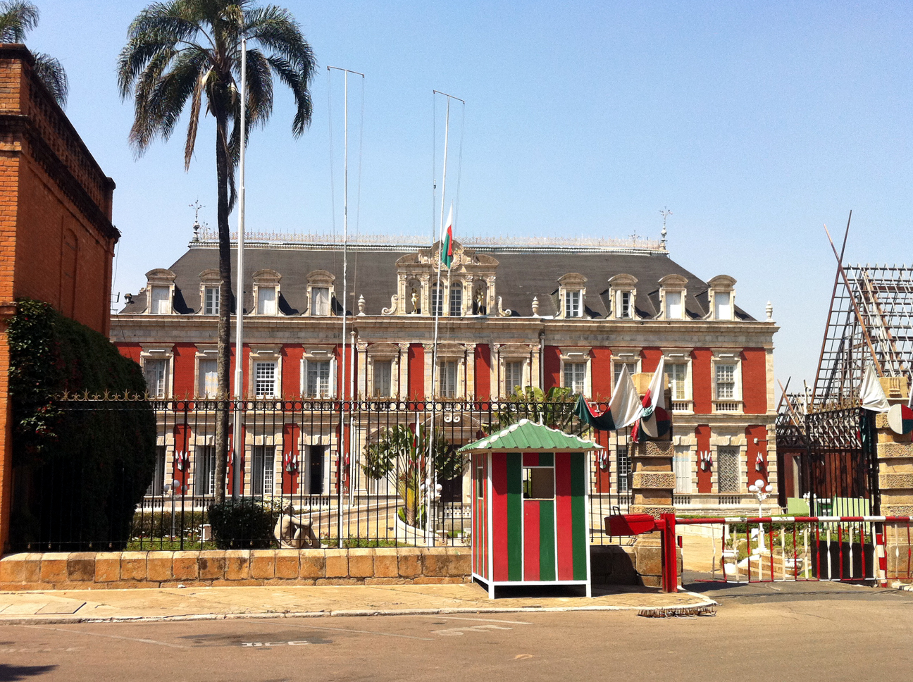 The red, white, and green Ambohitsorohita Palace in Antananarivo, Madagascar. The palace is shown from across the street, behind black metal railings, and a tall palm tree.