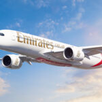 A white, red, and green Emirates jet Flying in front of a sunny sky. Some clouds are shown in the background.