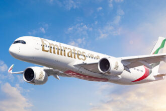 A white, red, and green Emirates jet Flying in front of a sunny sky. Some clouds are shown in the background.