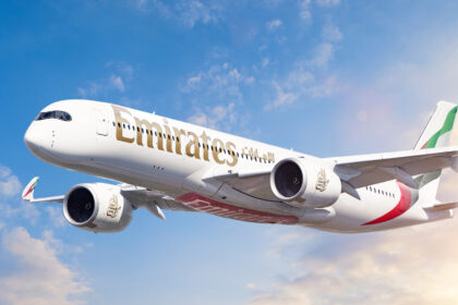 A white, red, and green Emirates jet Flying in front of a sunny sky. Some clouds are shown in the background.