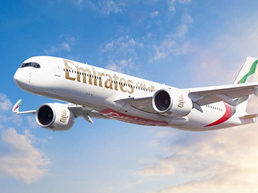 A white, red, and green Emirates jet Flying in front of a sunny sky. Some clouds are shown in the background.