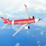 A red and white AirAsia jet flying over an ocean. A few clouds can be seen below the jet.