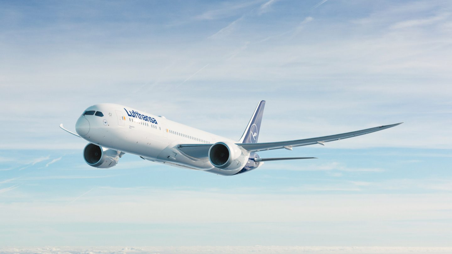 A white Boeing 787 jet painted in the livery of the Lufthansa Group. It is shown flying across a blue sky, obscured by a layer of cloud.