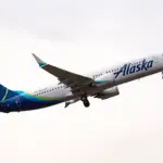 A blue and white Alaska Airlines B737-800 jet flying upwards in front of a cloudy sky.