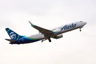 A blue and white Alaska Airlines B737-800 jet flying upwards in front of a cloudy sky.