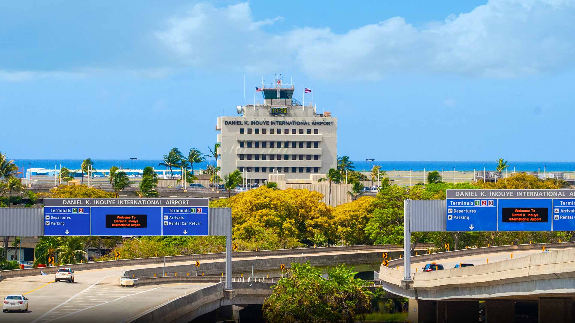 Daniel K. Inouye International Airport (HNL) in Honolulu, Hawaii. The airport terminal is shown behind a yellow and green bush, and in front of a clear blue sky.