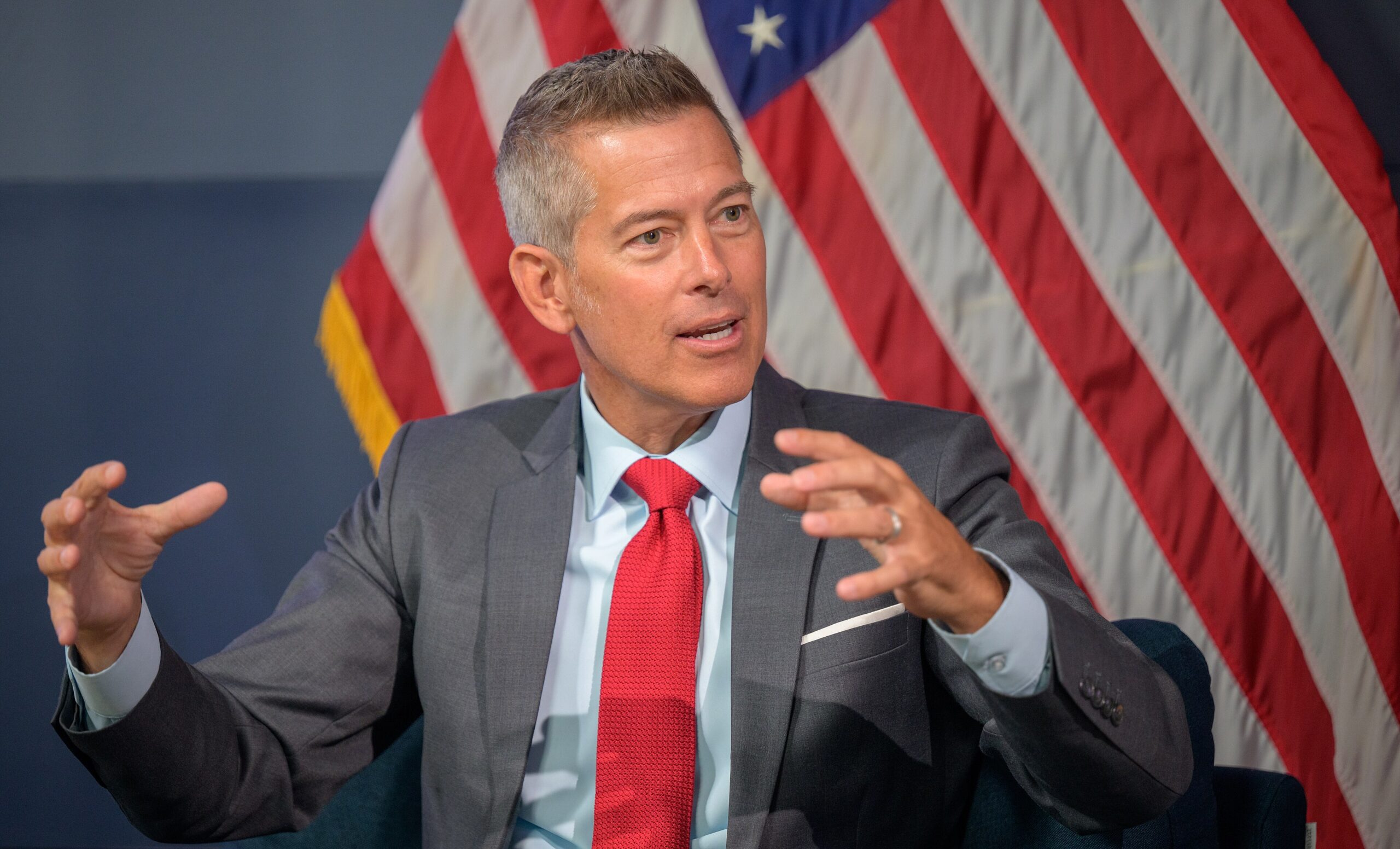 Sean Duffy speaking at a press conference. Duffy is sat in front of the American flag, and is wearing a grey suit with a red tie.
