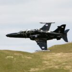 A black Royal Air Force RAF T2 Hawk flying over grassy hills in front of a cloudy sky.