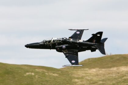 A black Royal Air Force RAF T2 Hawk flying over grassy hills in front of a cloudy sky.