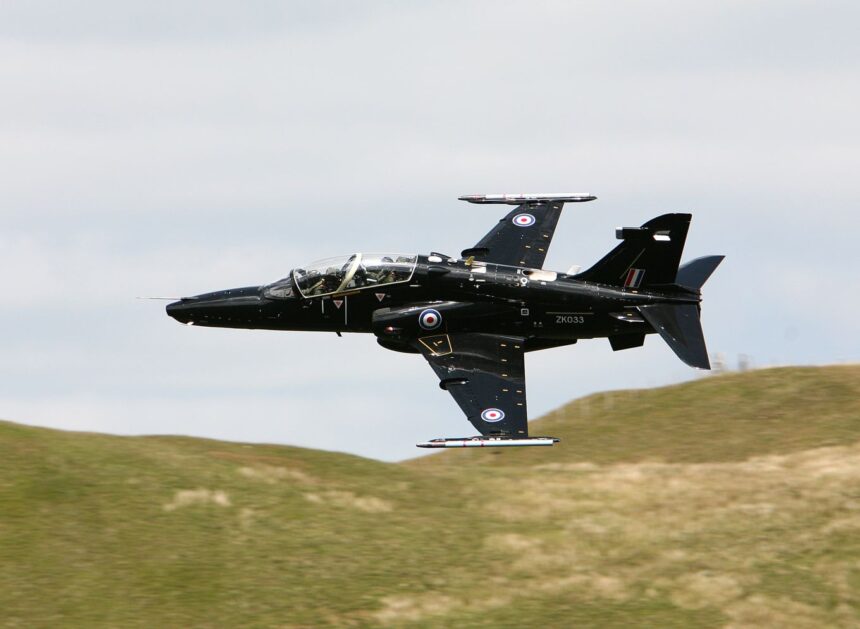 A black Royal Air Force RAF T2 Hawk flying over grassy hills in front of a cloudy sky.