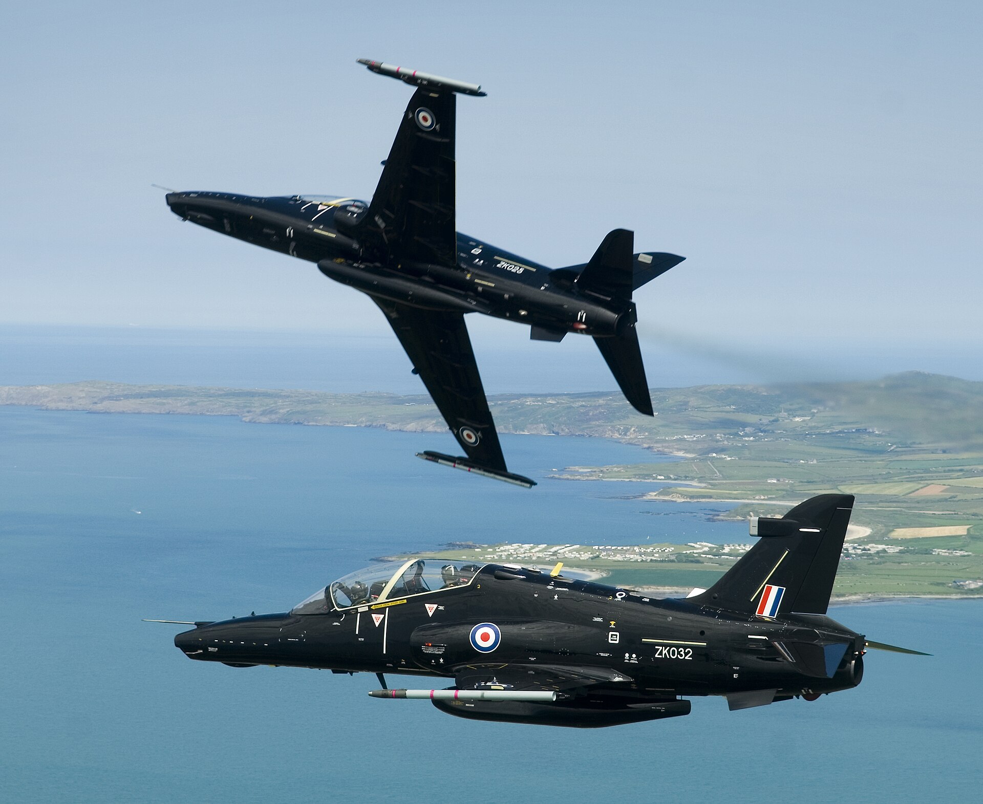 Two black RAF T2 Hawk planes flying over the North Wales coastline. One of the planes is beginning a barrel roll manoeuvre.