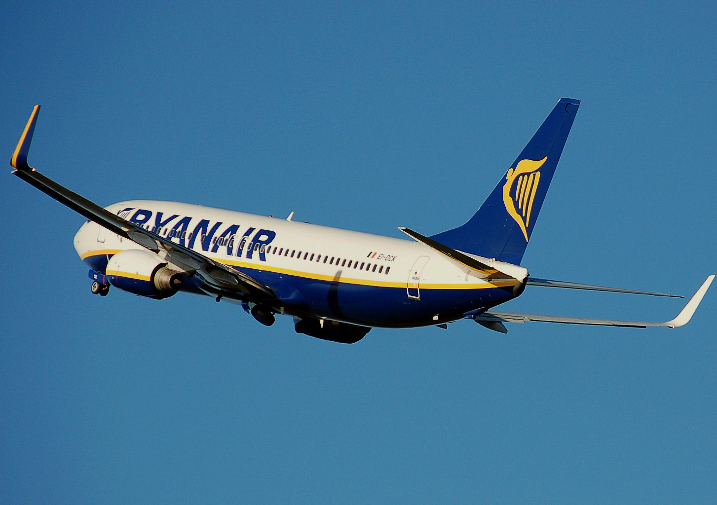 A blue, white, and yellow Ryanair jet flying in front of a clear blue sky.
