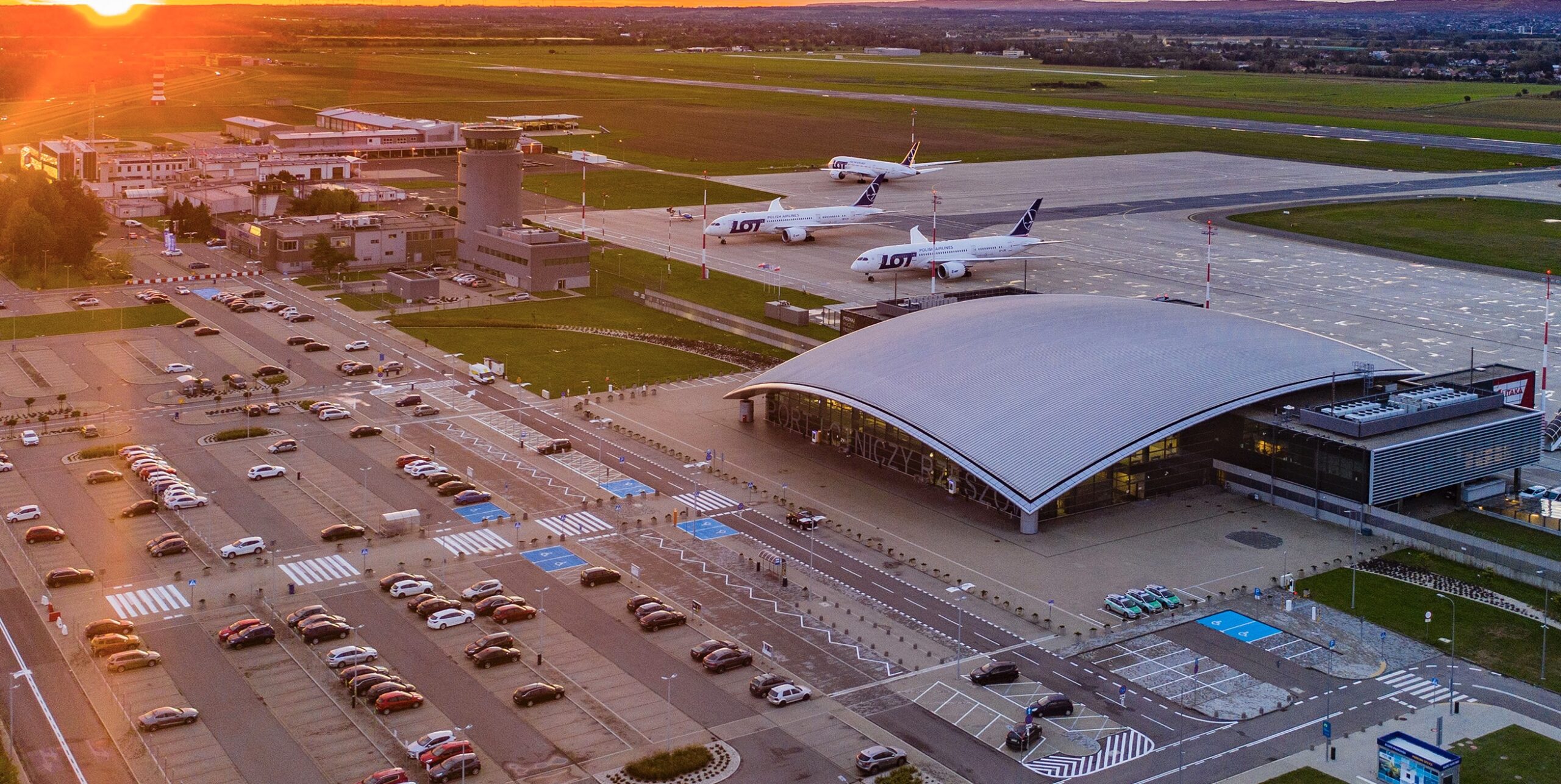 Sunset over RZE in Poland. A grey terminal building is seen between a runway, where LOT Polish Airlines jets are sat, and a car park.