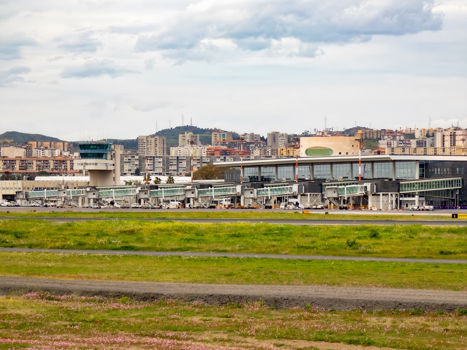 The outside exterior view of Catania-Fontanarossa Airport in Italy, featuring glass windows, buildings behind, and a runway at the front. 
