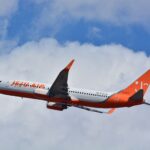 A Jeju Air Boeing 737-800 is pictured in flight from underneath. The aircraft is mostly white with orange branding on the side of the fuselage, as well as on the backend and tail. The aircraft is in the centre of the image, with the nose facing left and the tail on the right. The background is a bright blue sky with fluffy white clouds.