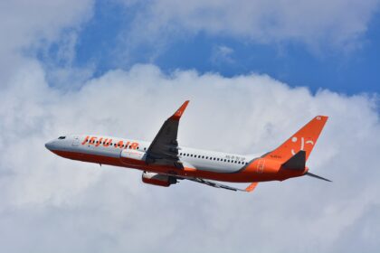 A Jeju Air Boeing 737-800 is pictured in flight from underneath. The aircraft is mostly white with orange branding on the side of the fuselage, as well as on the backend and tail. The aircraft is in the centre of the image, with the nose facing left and the tail on the right. The background is a bright blue sky with fluffy white clouds.