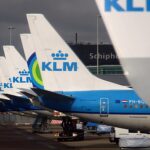 Several KLM aircraft tails are pictured lined up in a row at Schiphol Airport in Amsterdam, getting further away from the photographer. The tails are white, with mid blue underbellies and KLM branding on the tail in the centre.