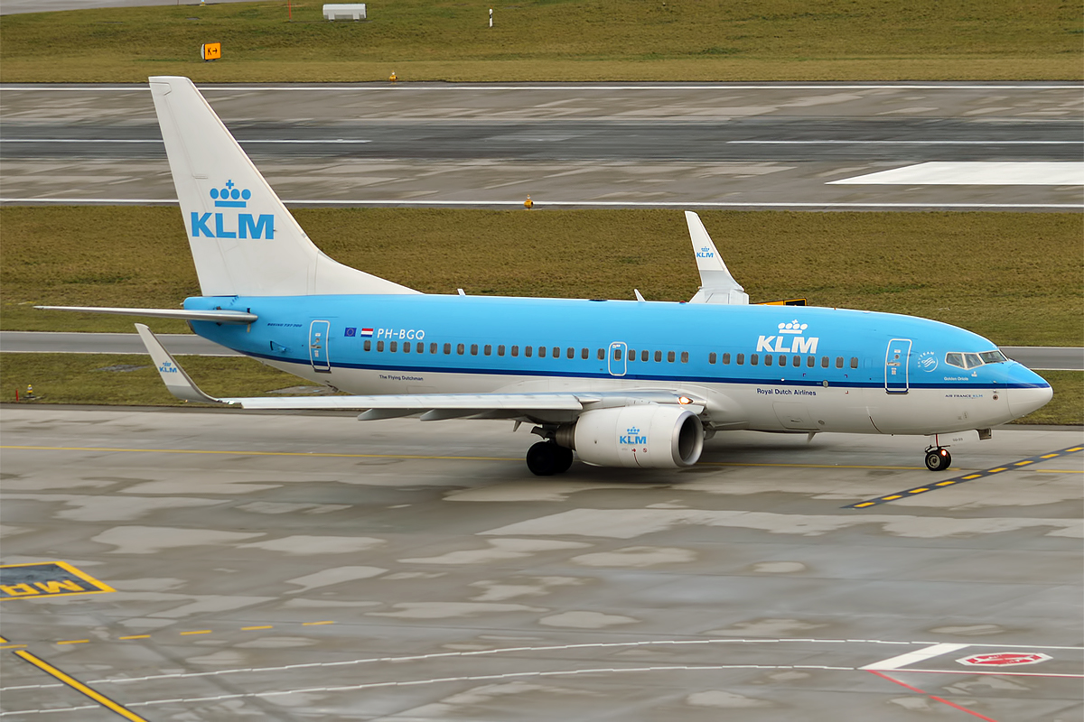 A KLM aircraft is pictured at an airport in a taxiway. The aircraft is pale blue with a white underbelly and tail. The tail, which is positioned in the left side of the image, has pale blue KLM branding in the centre, including the crown-like logo. In the background, grass verges separate a tarmac runway from the taxiway.