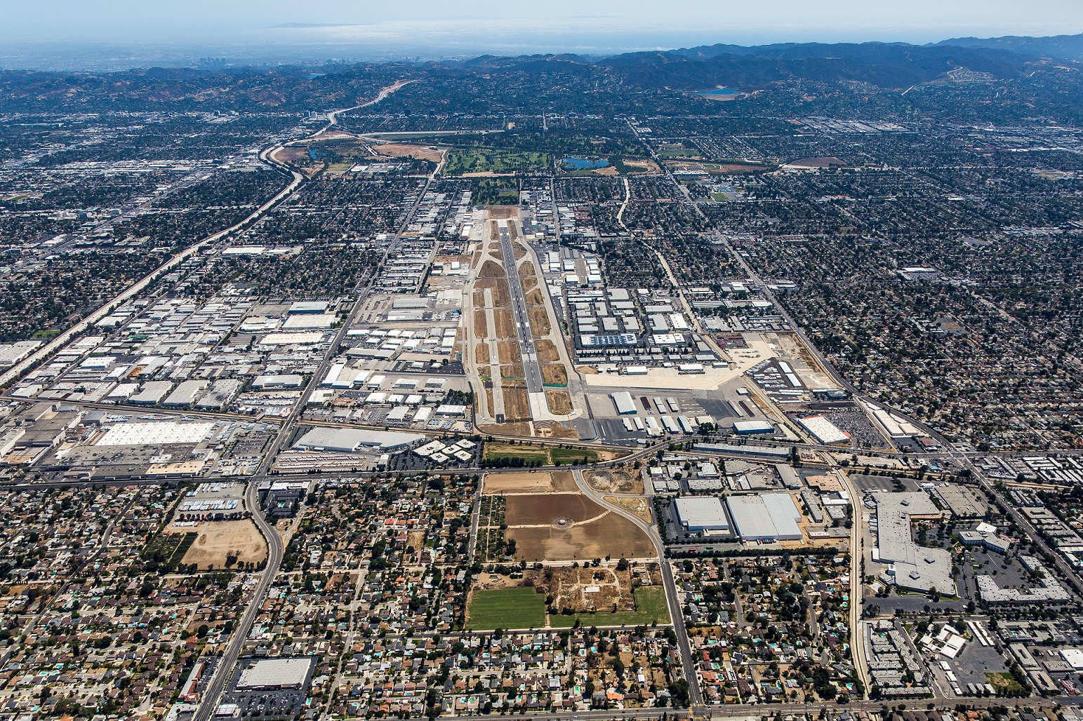 High-angle aerial view of an Van Nuys airport surrounded by a dense suburban layout, commercial buildings, roads, and distant mountains under a clear sky.