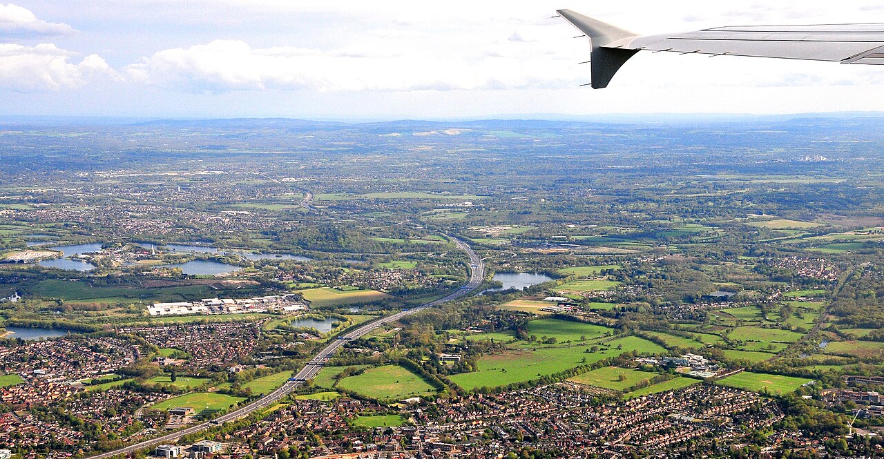 Looking southwards towards runway 27, to the left of London's Heathrow Airport, the M25 motorway can be clearly seen. The surrounding area is filled with vibrant greenery and dotted with visible reservoirs.