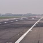 A runway at Heathrow Airport. The runway is grey tarmac, lined with lights. A white stripe travels down the centre. A grey sky can be seen in the background, along with grassland.