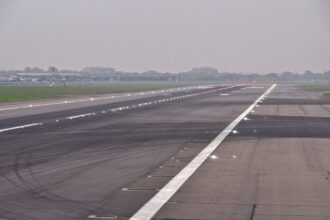 A runway at Heathrow Airport. The runway is grey tarmac, lined with lights. A white stripe travels down the centre. A grey sky can be seen in the background, along with grassland.