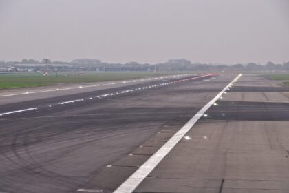 A runway at Heathrow Airport. The runway is grey tarmac, lined with lights. A white stripe travels down the centre. A grey sky can be seen in the background, along with grassland.