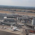 A part of London's Heathrow International Airport with a couple planes stationed around. The grass around the runway is dry and yellow. The airport has an air of busyness despite it not being entirely visible.