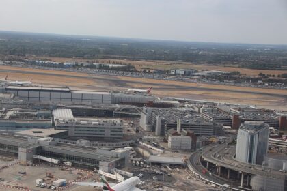 A part of London's Heathrow International Airport with a couple planes stationed around. The grass around the runway is dry and yellow. The airport has an air of busyness despite it not being entirely visible.