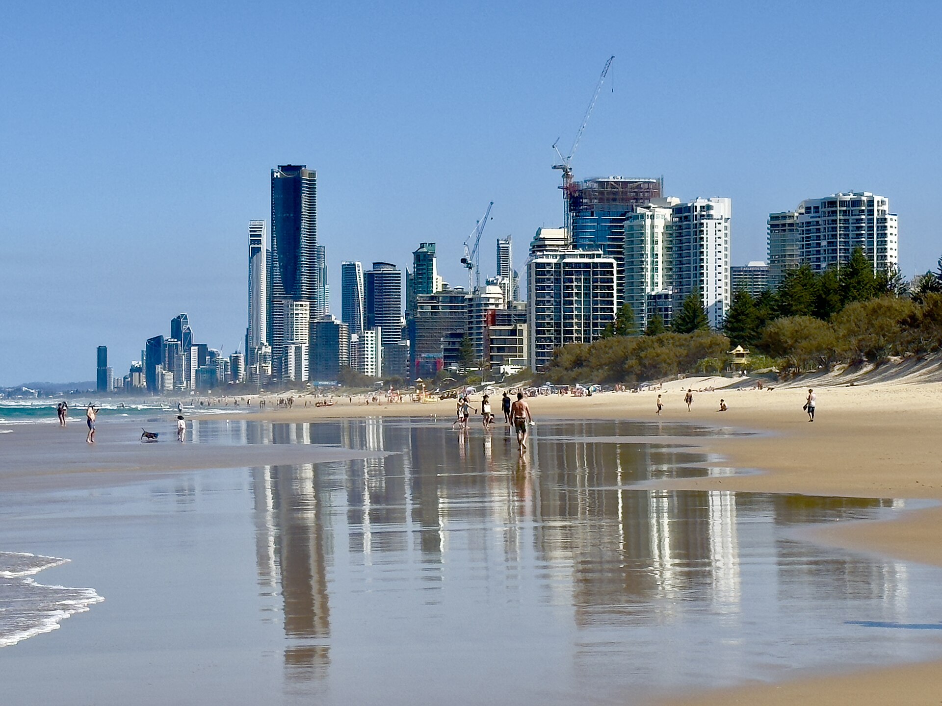 A large beach along the Gold Coast in Queensland Australia is pictured in September 2023. The sea is just about visible to the left. The foreground depicts sand and a layer of water from the tide having washed over it, and people walking. In the background, large buildings of the city can be seen against a bright blue cloudless sky. 