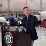 Sean Duffy speaking at a podium bearing the Federal Aviation Administration seal inside an aircraft hangar with planes visible in the background.