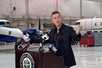 Sean Duffy speaking at a podium bearing the Federal Aviation Administration seal inside an aircraft hangar with planes visible in the background.