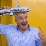 Ryanair CEO pictured against a yellow background, as he jokingly points upwards toward a model Ryanair aircraft, which he holds on top of his head.
