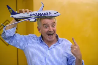 Ryanair CEO pictured against a yellow background, as he jokingly points upwards toward a model Ryanair aircraft, which he holds on top of his head.