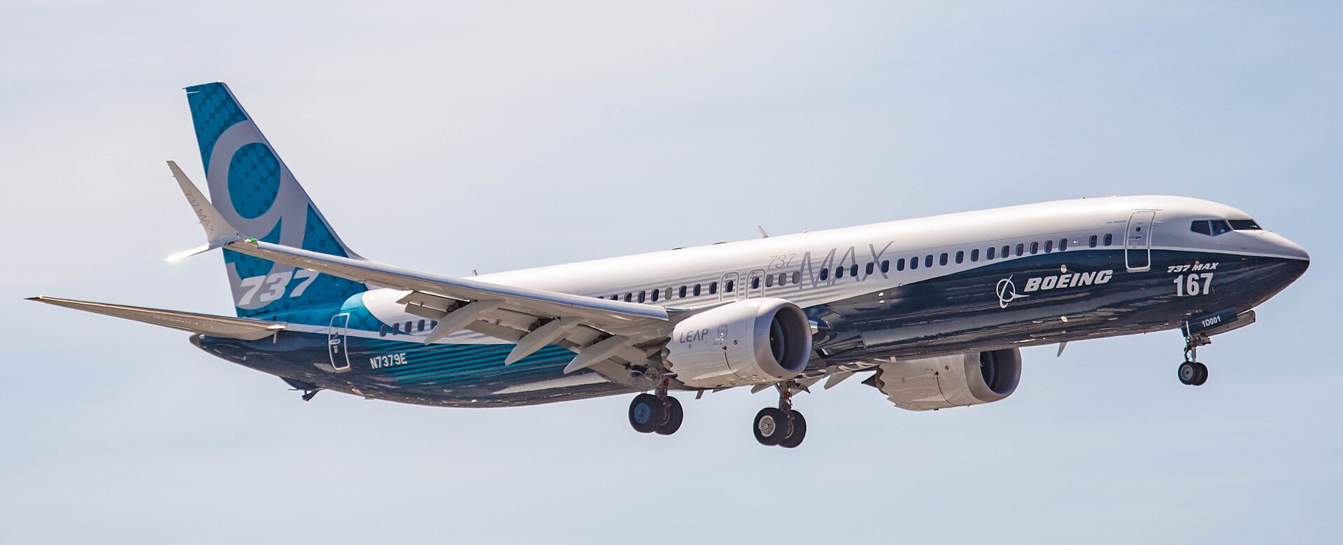 A Boeing 737 MAX 9 aircraft is pictured in flight. The aircraft is mostly white, with dark navy blue underbelly towards the front of the plane, and a mid blue backend and tail. The model type is stylistically printed in white on the tail. The aircraft is pictured at a slight angle, with the nose facing to the right and positioned upper centre far right and the tail positioned lower centre left. Pale cloudy sky occupies the background.