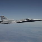 A NASA X-59 aircraft is pictured in flight along the Bell X 1 Supersonic Corridor in California. The aircraft has the sharp sleek edges typical r supersonic vehicle, and is white with NASA branding on the tail. The background is a clear muted blue sky with some clouds.