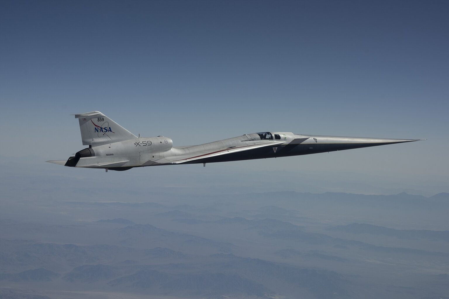 A NASA X-59 aircraft is pictured in flight along the Bell X 1 Supersonic Corridor in California. The aircraft has the sharp sleek edges typical r supersonic vehicle, and is white with NASA branding on the tail. The background is a clear muted blue sky with some clouds.