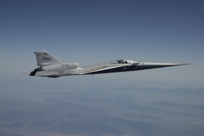 A NASA X-59 aircraft is pictured in flight along the Bell X 1 Supersonic Corridor in California. The aircraft has the sharp sleek edges typical r supersonic vehicle, and is white with NASA branding on the tail. The background is a clear muted blue sky with some clouds.