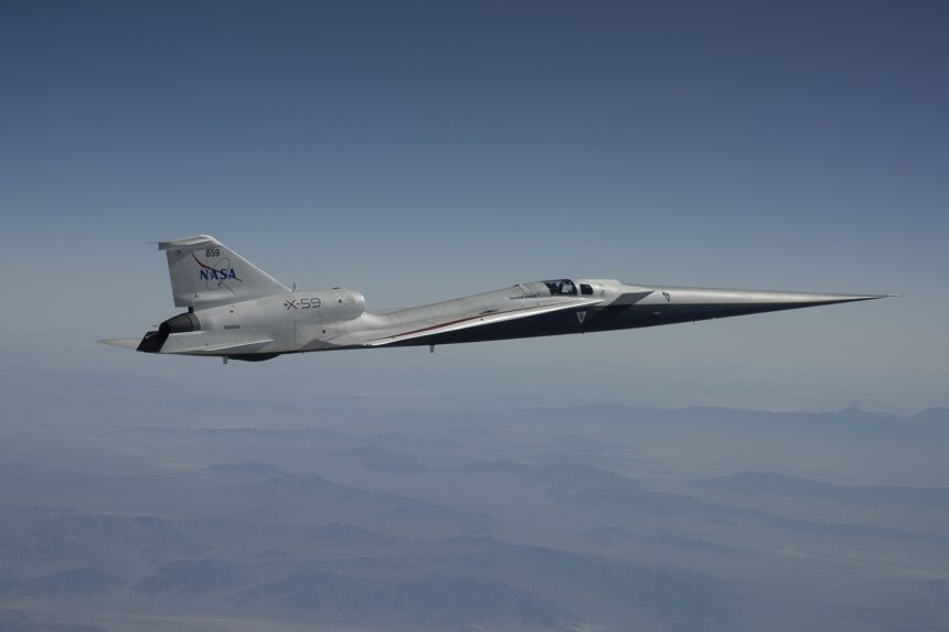 A NASA X-59 aircraft is pictured in flight along the Bell X 1 Supersonic Corridor in California. The aircraft has the sharp sleek edges typical r supersonic vehicle, and is white with NASA branding on the tail. The background is a clear muted blue sky with some clouds.