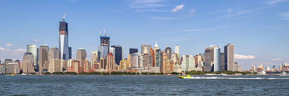 Image shows a view of the Manhattan skyline in New York City during the day with skyscrapers, water and a boat visible