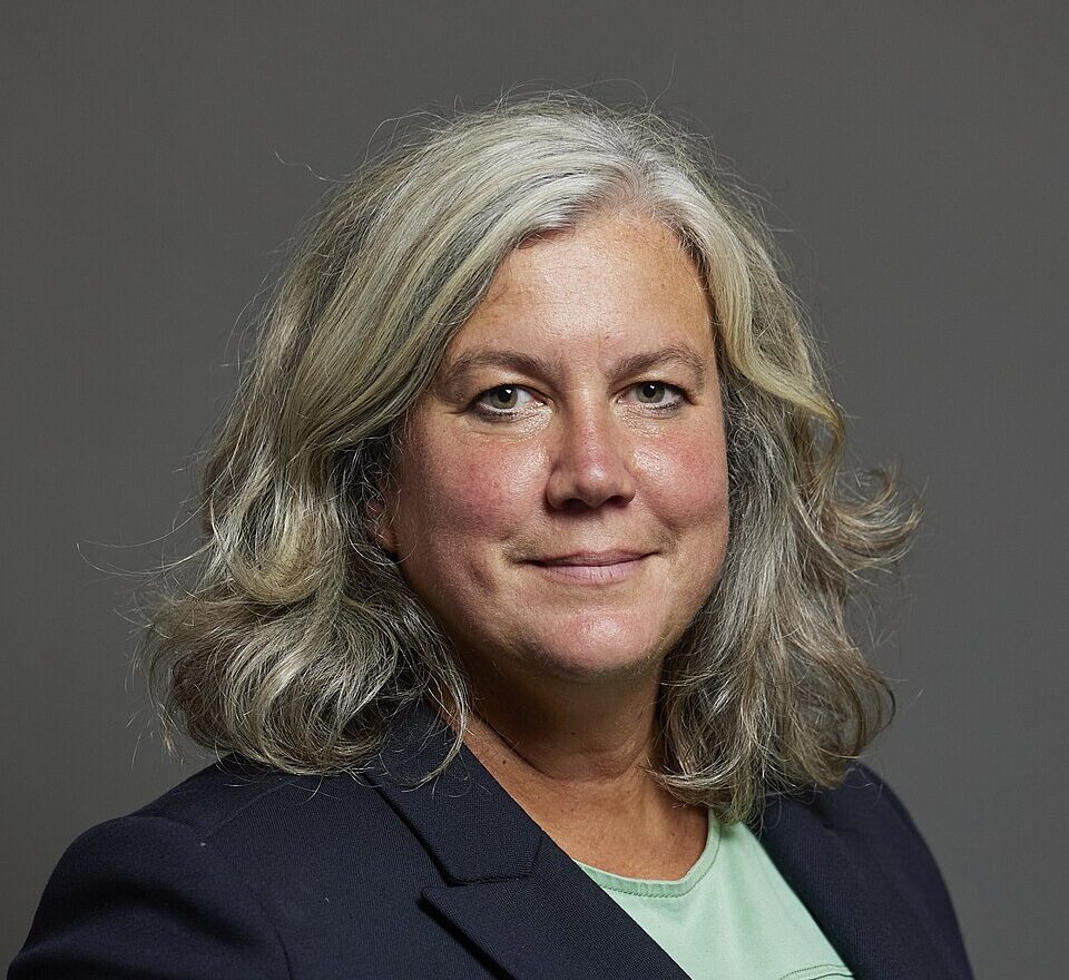 The official portrait of Transport Secretary Heidi Alexander. Alexander is a middle aged Caucasian female presenting individual with shoulder length platinum greyish hair and green eyes. She is wearing a navy blue suit jacket and a teal coloured top underneath. She is smiling close-mouthed, and has a dimple under her lip on one side. The background is grey. 