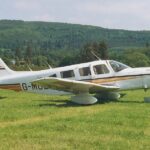 Image shows a Piper Cherokee 6 with yellow and white livery. It's parked on grass with a hill in the background.