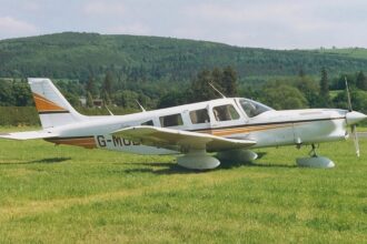 Image shows a Piper Cherokee 6 with yellow and white livery. It's parked on grass with a hill in the background.