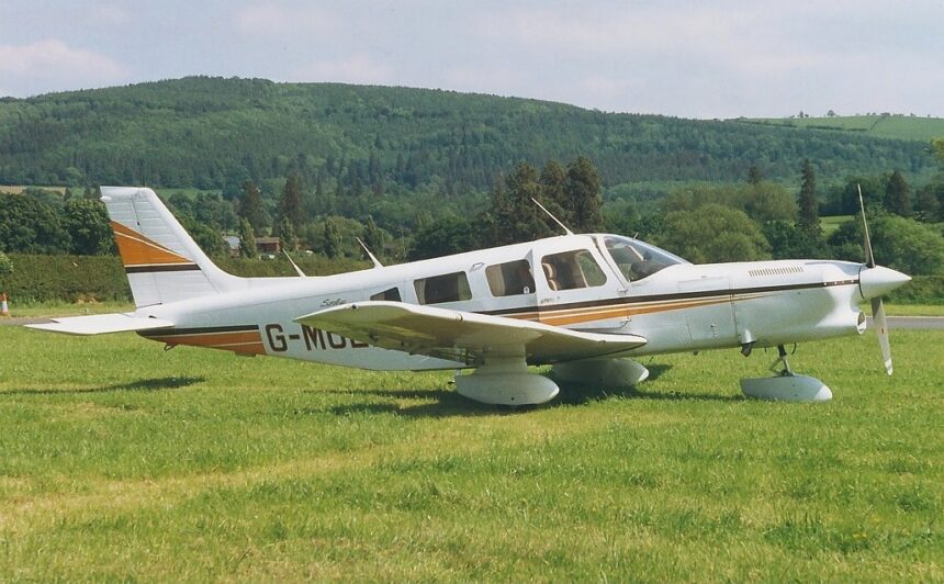 Image shows a Piper Cherokee 6 with yellow and white livery. It's parked on grass with a hill in the background.