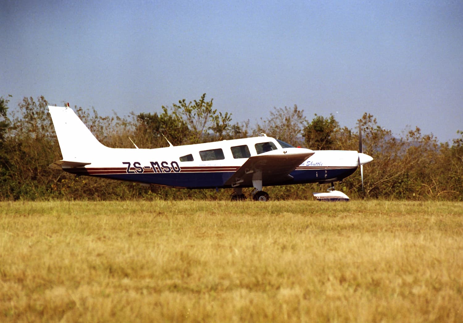 Image shows a Piper Cherokee 6 with white, blue and red livery. It's parked on grass with trees in the background.