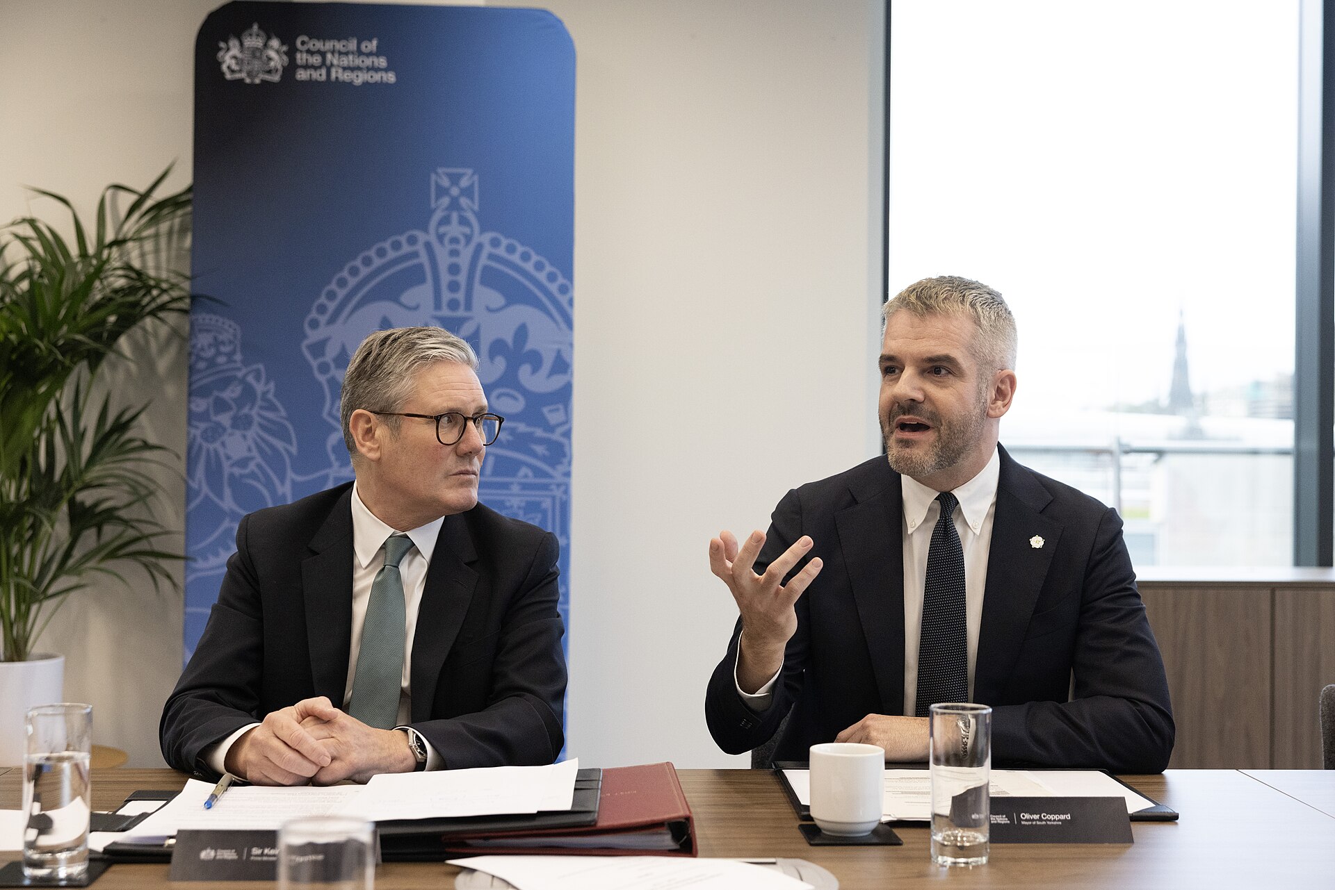 South Yorkshire Mayor Oliver Coppard (right) and Sir Keir Starmer (left) sit beside one another at a brown wooden table, with folders, documentation, and a glass of water in front of each of them. They are sat in front of a blue banner that reads 'Council of the Nations and Regions' beside the government crest. Both are dressed in dark suits with white shirts, and Coppard wears a dark tie and Starmer wearing a light bluish grey tie. Coppard is gesticulating with his hand and appears to be mid sentence, and Starmer looks at him.