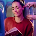 A flight attendant adjusts the flexible leather headrest for a passenger reading an open book under soft cabin lighting.