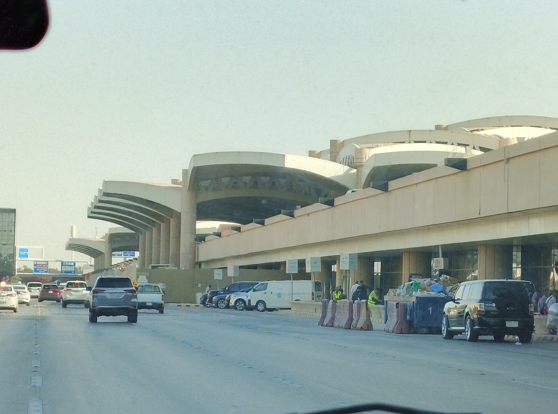 External view of King Khaled International Airport in Riyadh. The image is taken from an angle across the road, potentially from inside a vehicle on the road. The sand collared walls and pillars of the structure occupy the majority of the left side, disappearing into the distance as the structure approaches the centre. Most of the foreground depicts concrete road, while the background depicts greyish blue sky. 