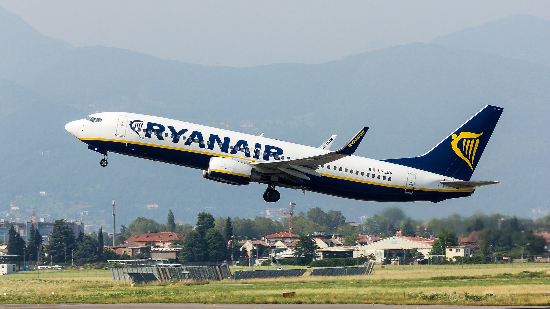 A Boeing 737-800 operated by budget airline Ryanair is pictured taking off. The aircraft is mostly white with a navy blue underbelly and tail, which has the deep yellow-gold logo on it. There is also navy blue lettered branding along the side, with a yellow stripe. The background is a greyish sky with mountains visible in the distance. Some residential properties can be seen just behind the airport boundaries. 