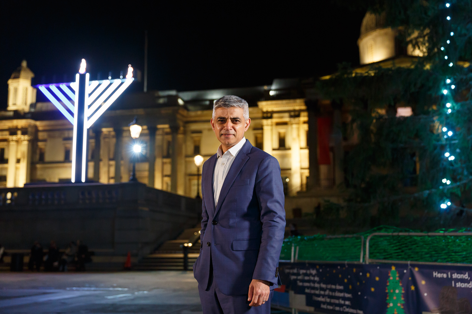 Mayor of London Sadiq Khan, stood in front of a public menorah placed by Chabad-Lubavitch at Trafalgar Square in London, England on December 10, 2020, on the first night of the eight-day holiday. The Mayor is wearing a smart blue suit with a white shirt under the buttoned jacket. The image is taken in the evening.